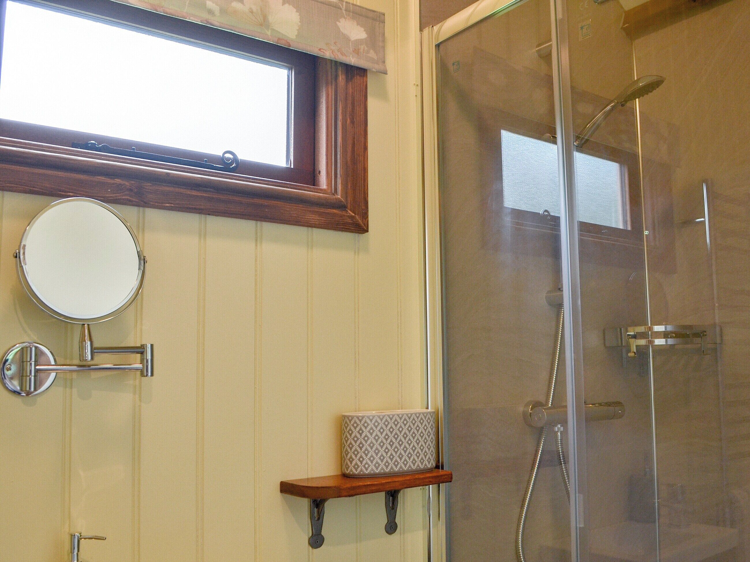 A bathroom with a glass-enclosed shower, a round mirror, and a small shelf with a patterned mug.