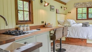 Een gezellige keuken met een houten aanrecht, een gasfornuis en een gootsteen. Er staat een bed met een witte dekbedovertrek en kussens, en een raam met uitzicht op groen.