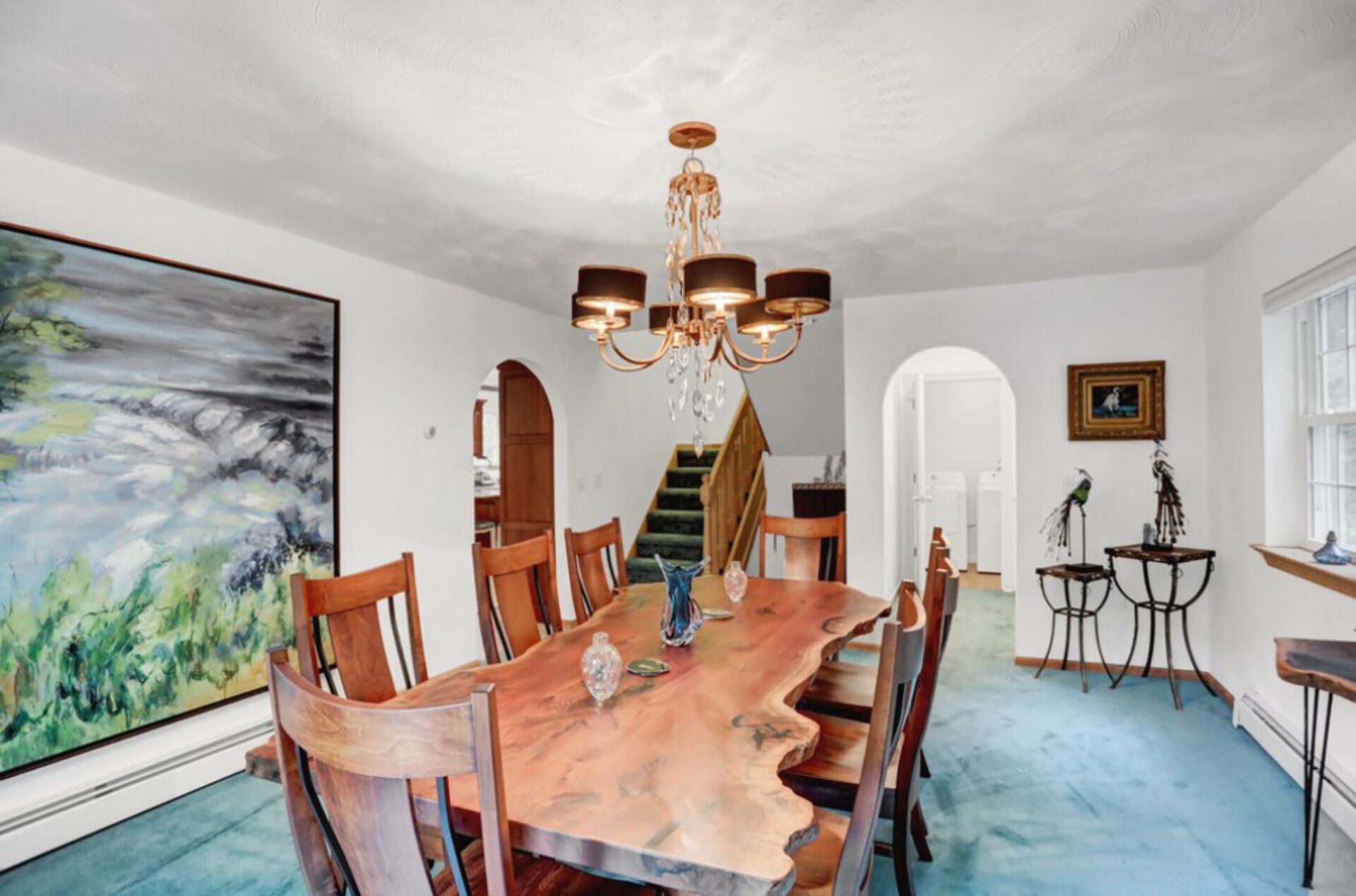 A dining room with a large wooden table, six chairs, a chandelier, and a large abstract painting on the wall.