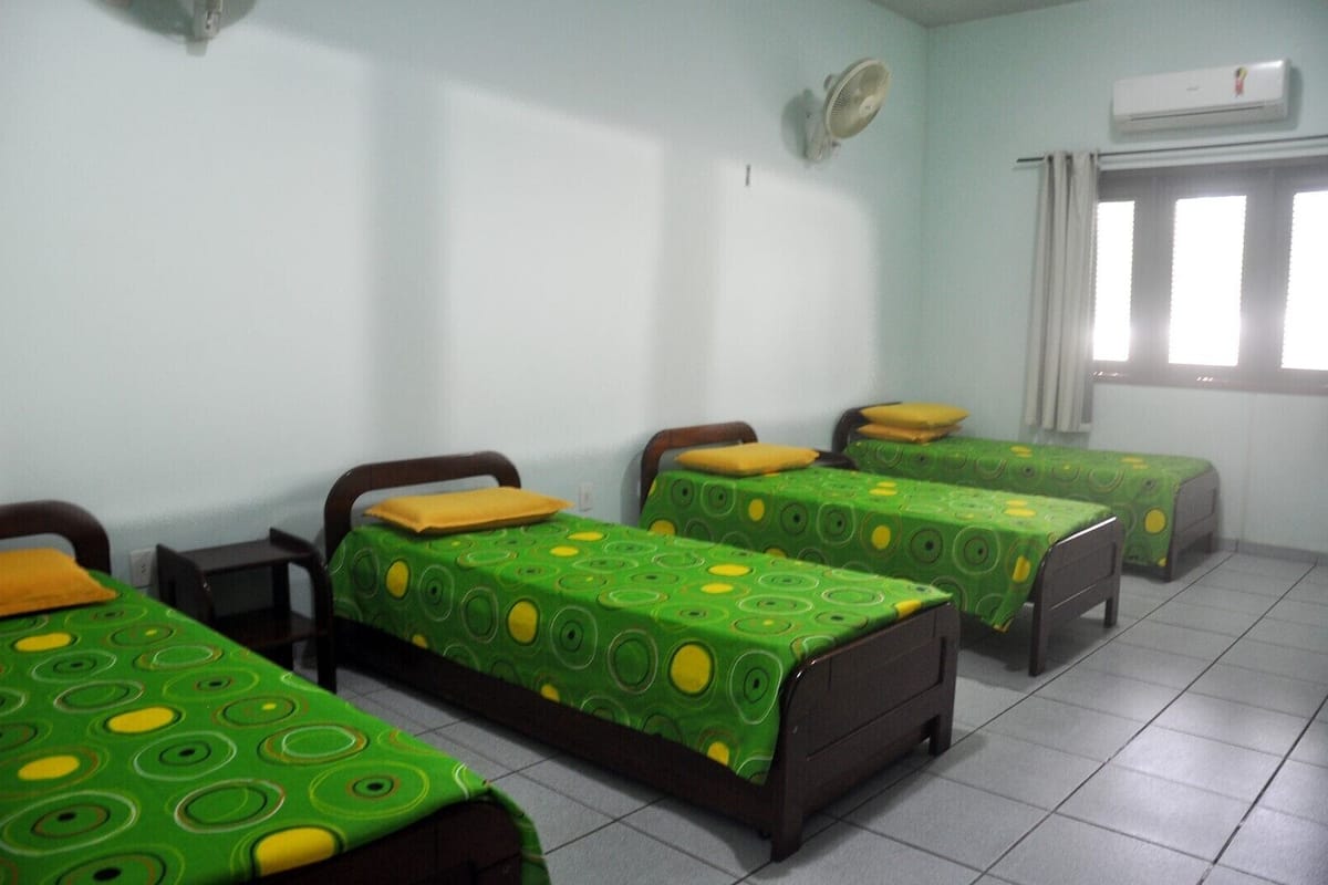 A dormitory room with four single beds, each with a green patterned bedspread and a yellow pillow. There is a window with curtains, an air conditioning unit on the wall, and a tiled floor.