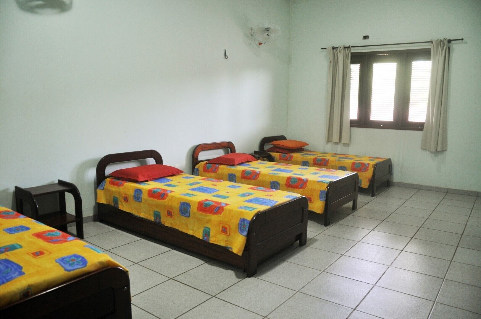 Una habitación con cuatro camas individuales, cada una con una colcha de colores, una silla y una ventana con cortinas.