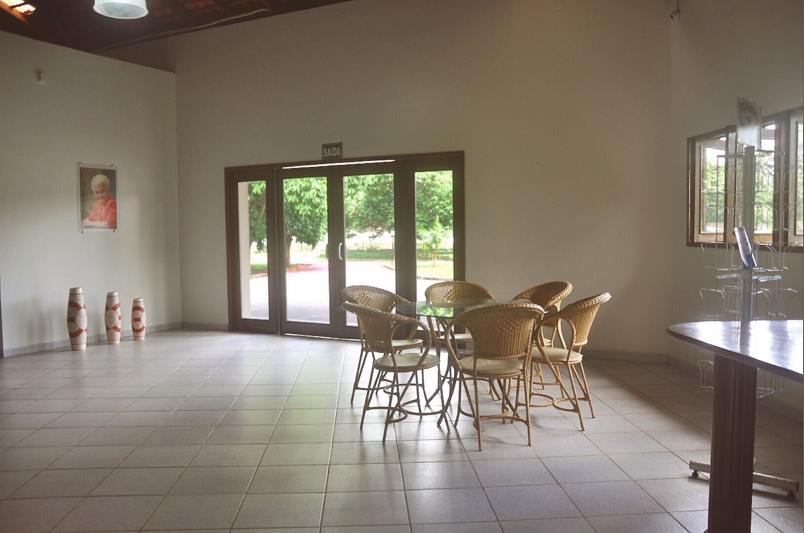 Una habitación amplia con un gran ventanal corredero que da a un espacio exterior, sillas de mimbre dispuestas alrededor de una mesa, y un cuadro en la pared.