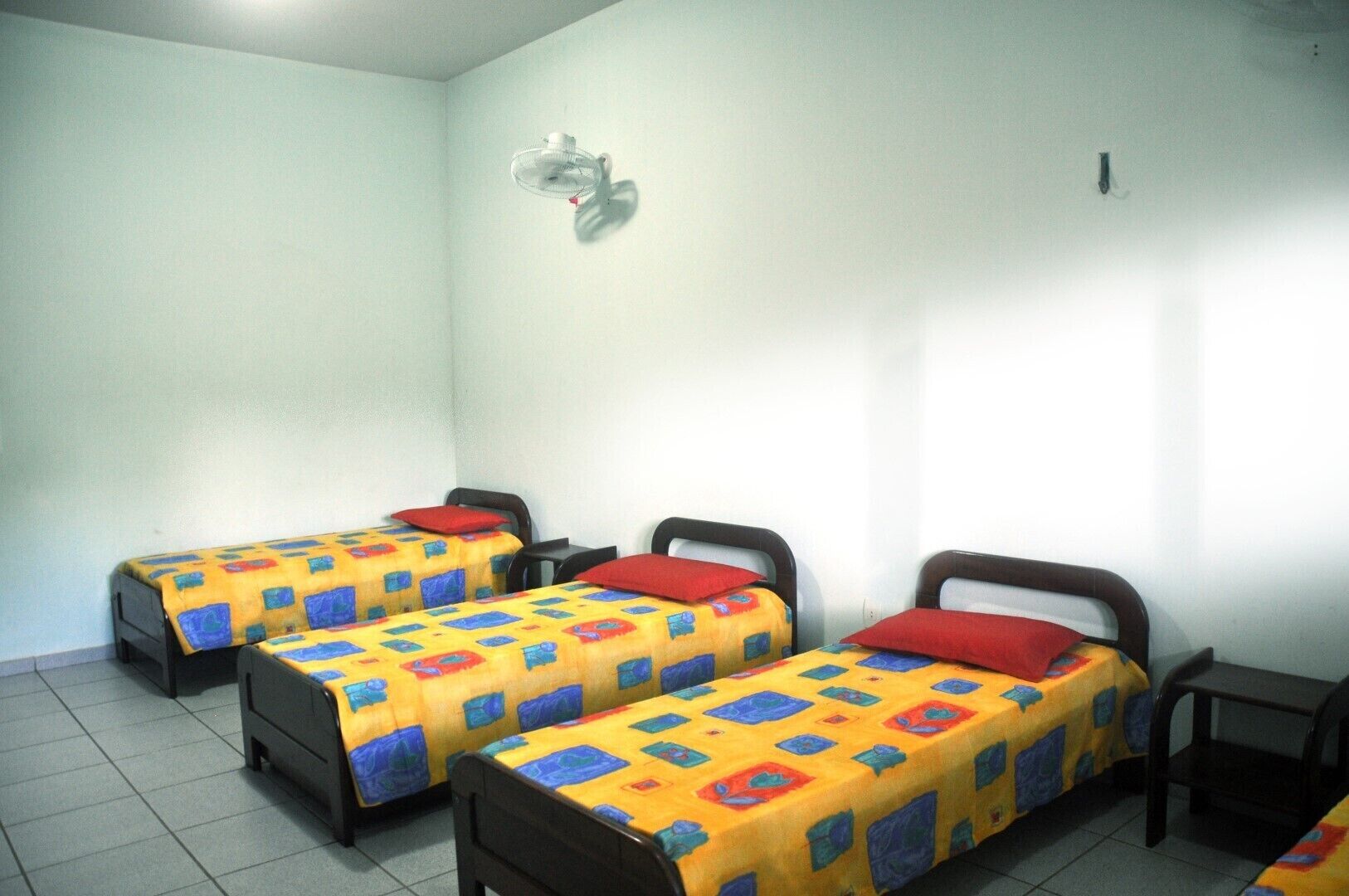 Una habitación con tres camas individuales, cada una con una colcha de colores y almohadas rojas, y una pequeña mesa entre las camas.
