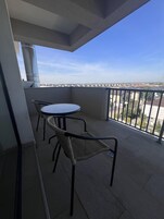 Un balcón con una mesa redonda y dos sillas de mimbre, vistas despejadas de la ciudad y una barandilla de metal.