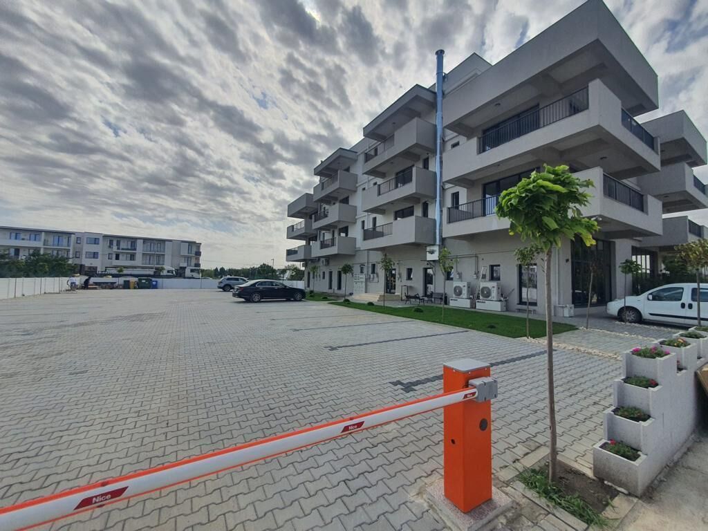 A modern multi-story building with balconies, a paved parking area, and a gated entrance.