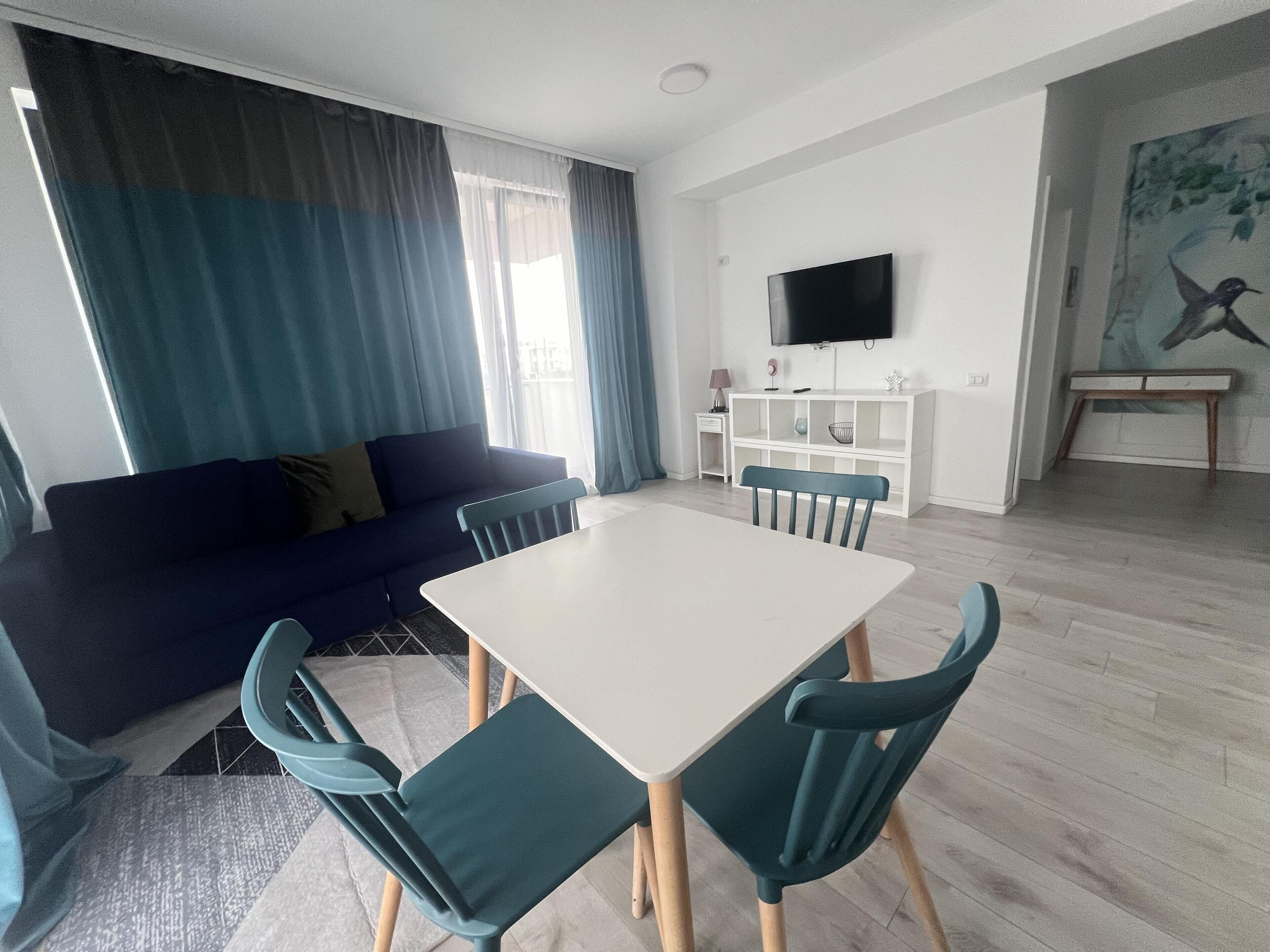 A modern living room with a sofa, a dining table, and chairs. There is a TV mounted on the wall and a painting on the wall.
