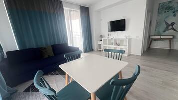 A modern living room with a sofa, a dining table, and chairs. There is a TV mounted on the wall and a painting on the wall.