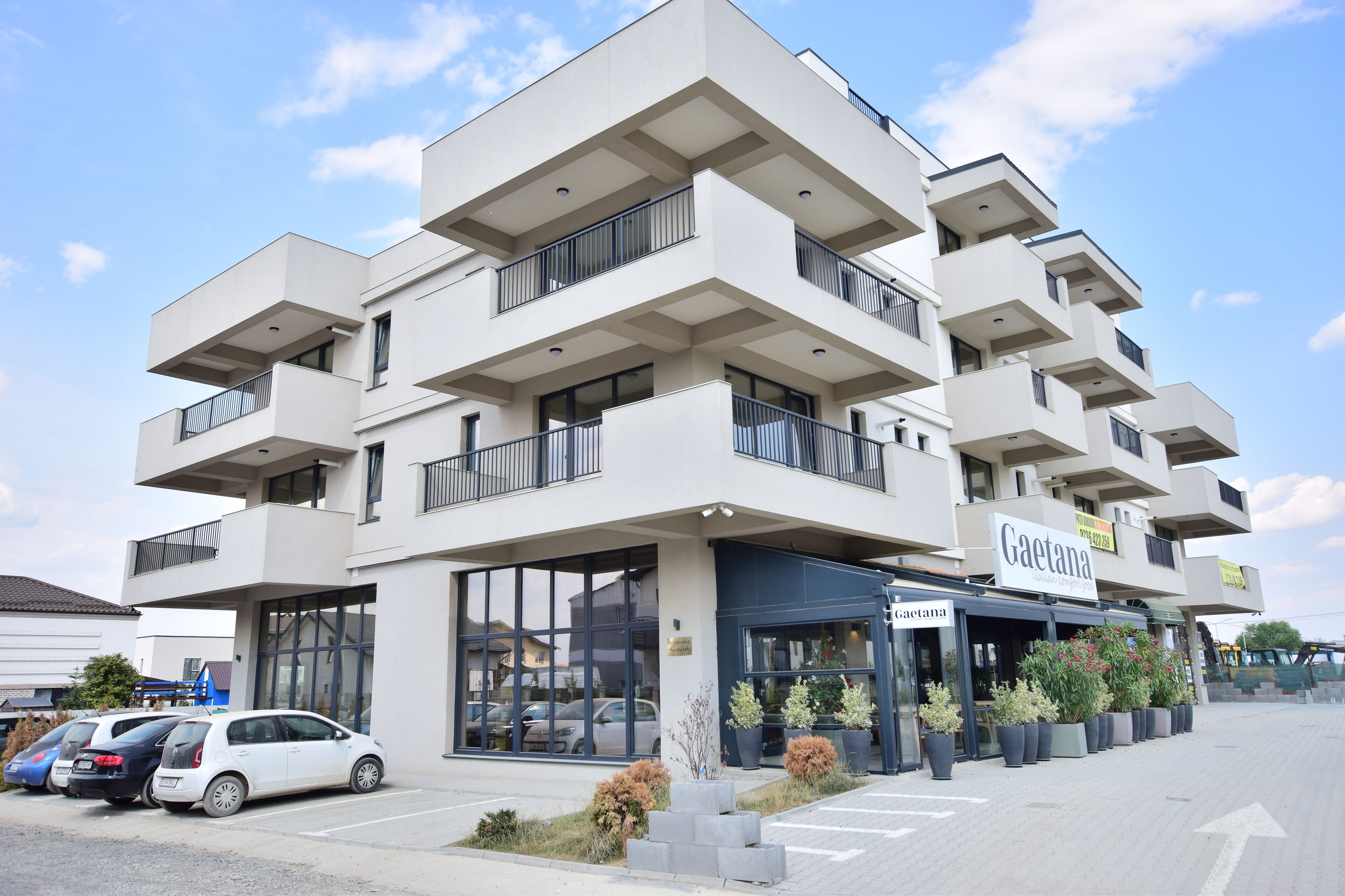 A modern multi-story building with a cafe named "Gaetanana" on the ground floor. There are several cars parked in front, and the building has balconies with railings.
