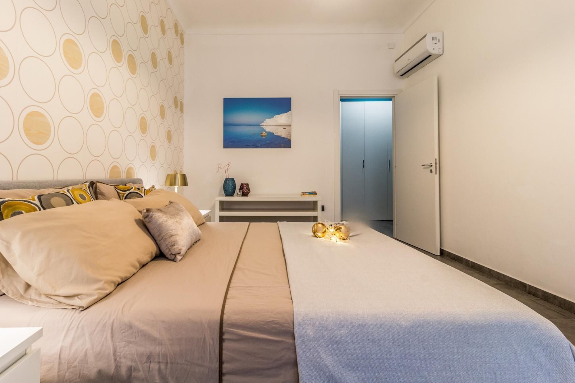 A bedroom with a bed, a nightstand, a painting, and an air conditioning unit.