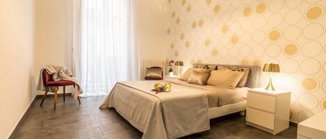 Un dormitorio moderno con cama, mesitas de noche, una silla y un amplio ventanal con cortinas traslúcidas.