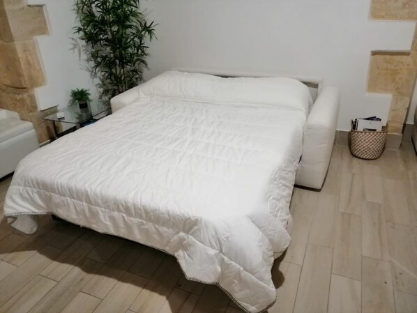 Una cama bien tendida con una colcha blanca en una habitación con suelo de baldosas, un sofá blanco, una mesita con tapa de vidrio y una planta en maceta.