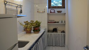 A compact kitchen with white tiles, a window with a view, and various kitchen items on shelves. - Villa de Leva - Casa Della Nonna (Modica)