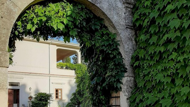 An arched stone gateway with ivy, leading to a building with a white facade and a balcony. - Villa de Leva - Casa Della Nonna (Modica)