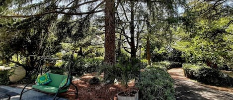Un espace extérieur paisible, aménagé avec un banc vert et une petite table, entouré d’arbres touffus et bordé d’un chemin de jardin bien entretenu.