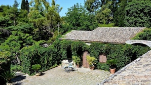 A stone building with a tiled roof, surrounded by lush greenery and a paved courtyard. - Villa de Leva - Casa Gialla (Modica)