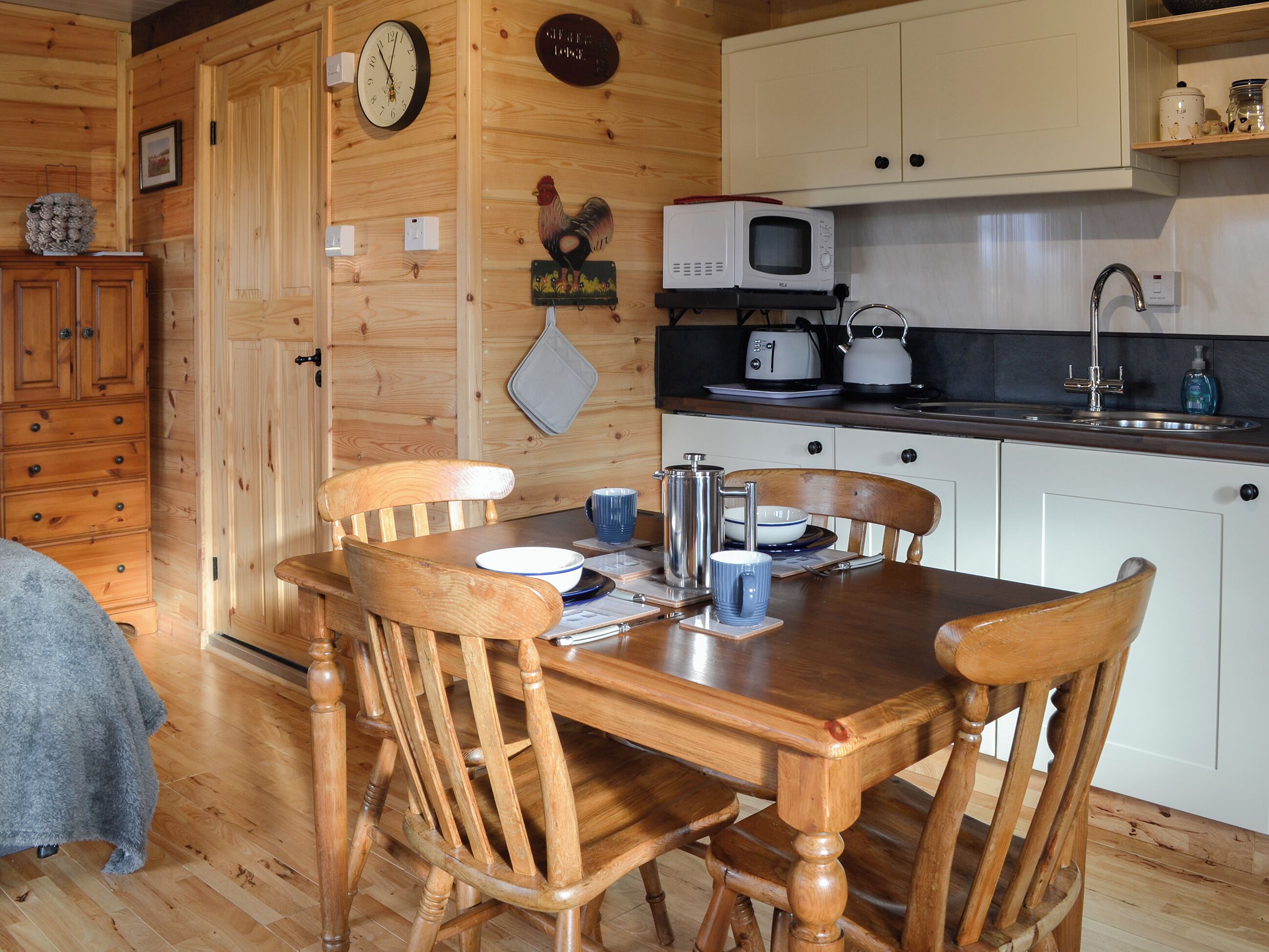 A wooden interior with a dining table, chairs, a microwave, a kettle, and a sink.
