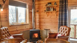 A cozy cabin interior with a wood-burning stove, leather chairs, a wooden table, and a bookshelf with books and decorative items.