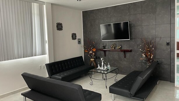 A modern living room with a black sofa, a glass coffee table, and a flat-screen TV mounted on a gray wall. - Casa a 5 Minutos del Hospital de La Mujer con Todos Los Servicios (Tehuacán)