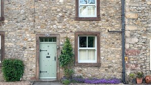 A stone building with a green door, two windows with shutters, and a small garden with purple flowers.
