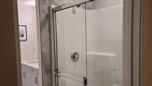 Une douche fermée par des parois de verre, avec un sol carrelé de carreaux blancs et un mur en marbre.