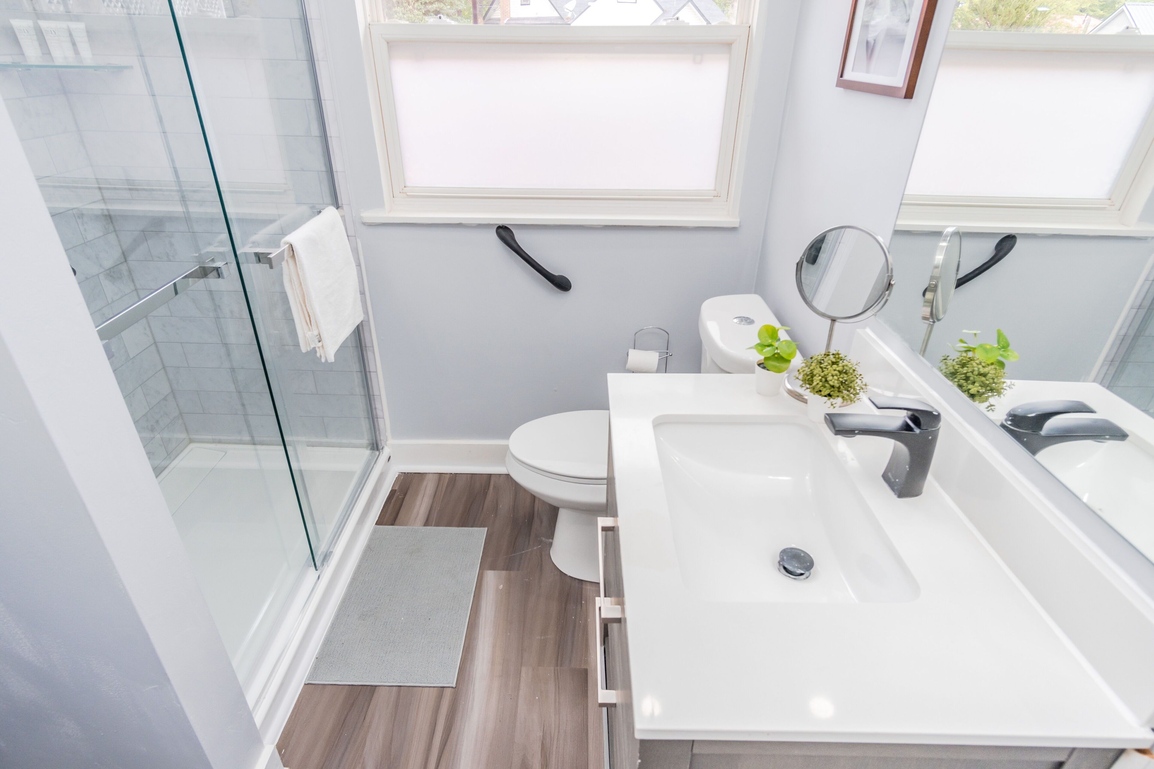A modern bathroom with a glass shower enclosure, a white sink with dual faucets, a toilet, and a small window above the sink.
