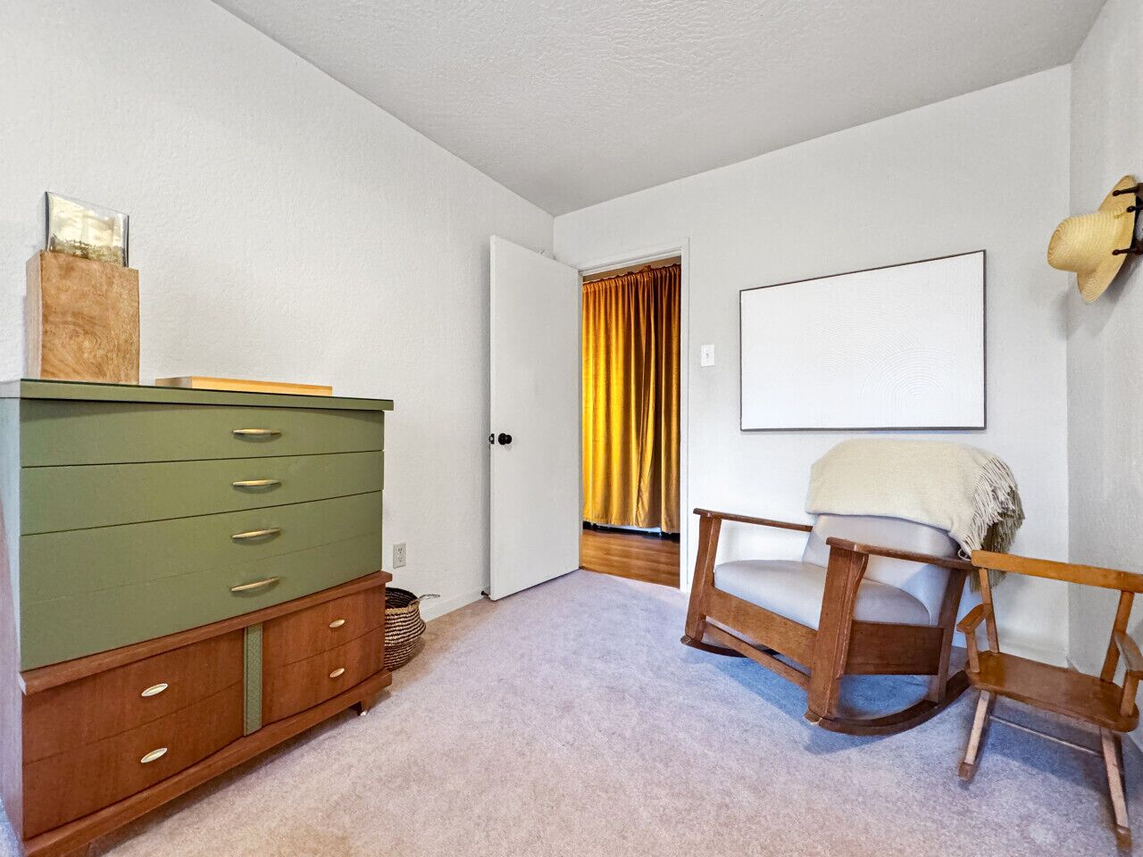 A room with a green dresser, a rocking chair, a framed picture on the wall, and a door with yellow curtains.