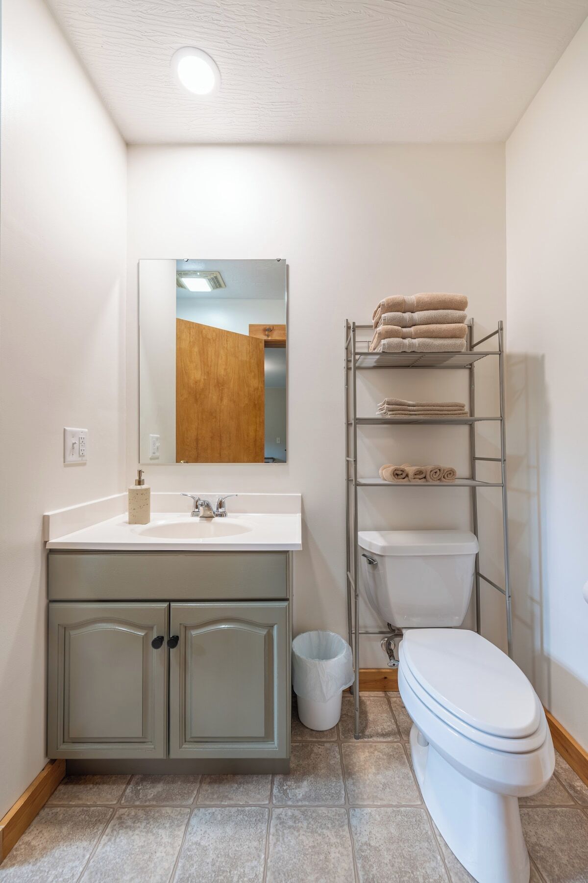 A bathroom with a white sink, mirror, and toilet. There is a towel rack with folded towels and a trash can.