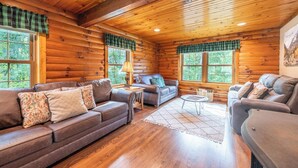 A log cabin interior with wooden walls, a brown leather sofa, a small round table, and a window with a view of trees.