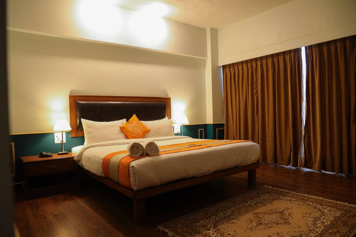 Habitación de hotel con cama de madera, ropa de cama blanca, un plaid naranja, una mesita de noche con lámpara y un ventanal con cortinas.