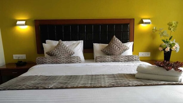 A hotel room with a large bed, two bedside tables, a nightstand, a wall-mounted lamp, and a vase with flowers. - Krishna Inn - The Green Hotel (Kolhapur)