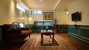 Un salón con un sofá rojo, una mesa de centro de madera y una alfombra estampada. Se observan dos ventanas, un cuadro en la pared y un televisor montado.