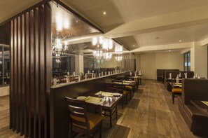 Un restaurant moderne doté d’un long comptoir, de lambris en bois et d’un lustre. Les tables sont disposées de manière ordonnée, avec des chaises.
