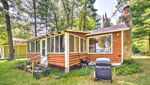 Une cabane en rondins avec un porche couvert, un barbecue et une poubelle, située dans une zone boisée.
