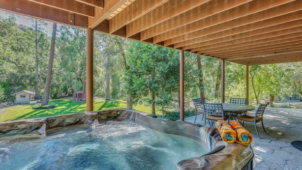 Un espace avec un jacuzzi, agrémenté d’une pergola en bois, d’une terrasse en pierre et offrant une vue sur une pelouse verdoyante et des arbres.