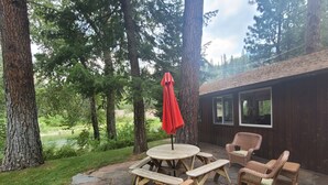 A patio area with a stone table and wooden chairs, surrounded by tall trees and a view of a river. - Fish, float, or raft out of your front door, MT's wilderness at your fingertips! (Bonner)