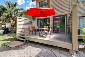 A covered patio with a table and chairs, a red umbrella, and a palm tree. - 3 Story Condo - Bonus Room Upstairs! (Hilton Head Island)