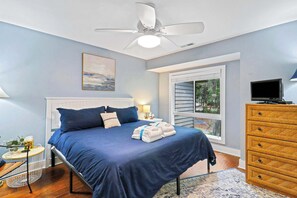 A bedroom with a bed, a ceiling fan, a window with blinds, a TV, a wooden dresser, and a nightstand with a lamp.