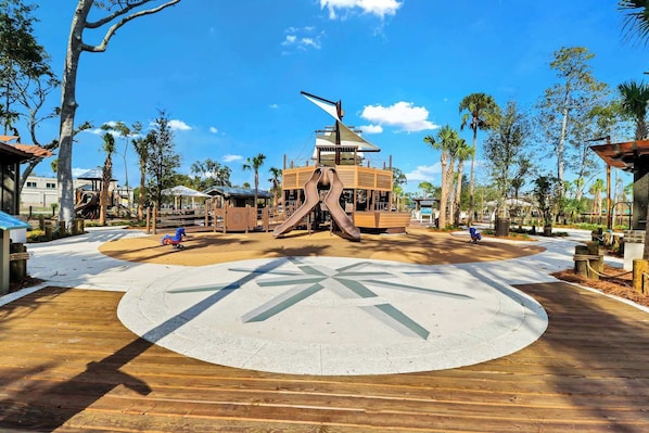 Un parque infantil con un tobogán central y estructuras de juego circundantes, con palmeras y cielos despejados de fondo.