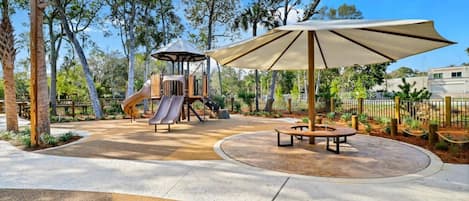 A playground with a slide and a play structure, surrounded by trees and a paved area with benches.