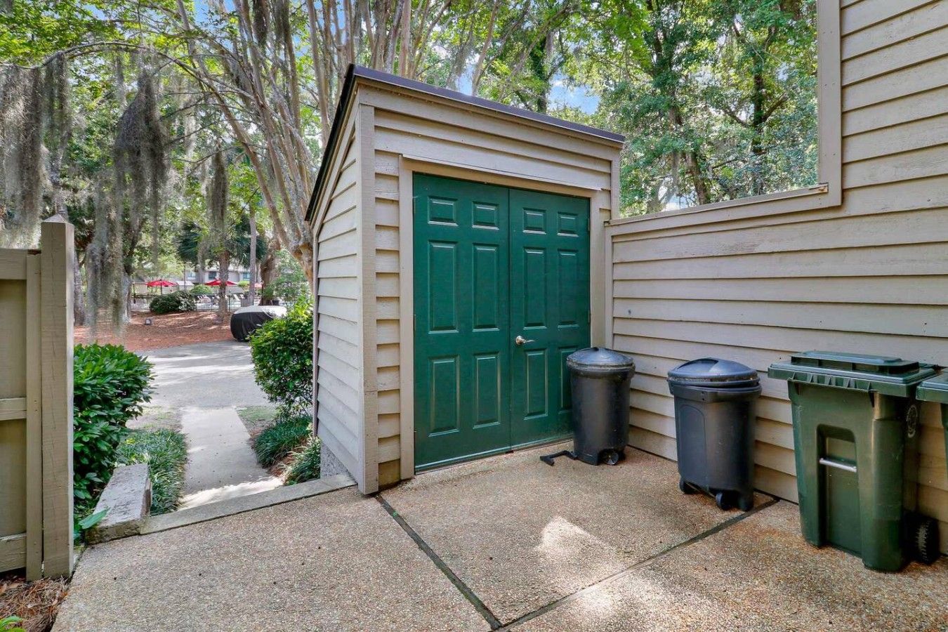 A small shed with a green door, two trash bins, and a paved pathway leading to a covered area.