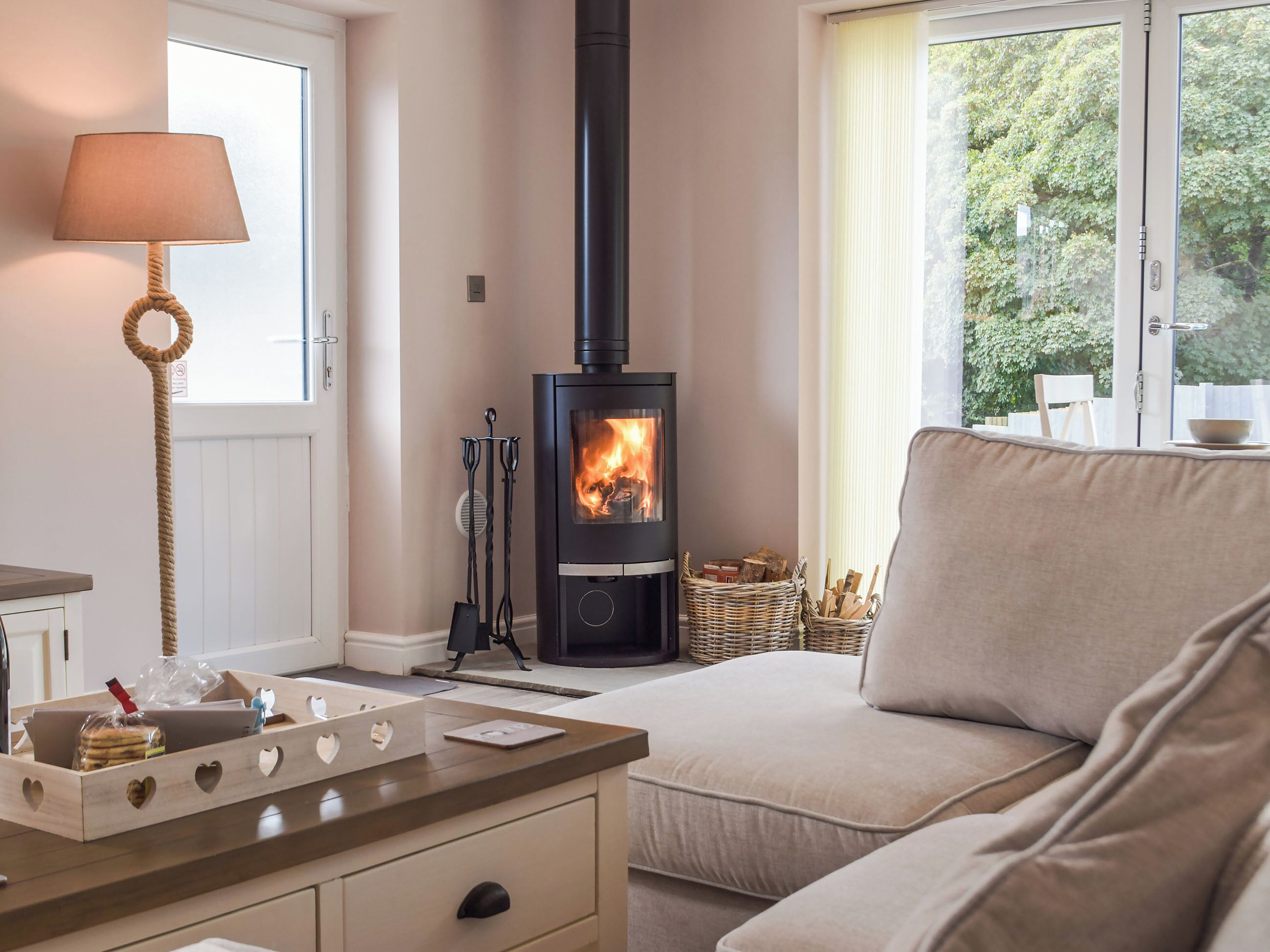 A cozy room with a sofa, a table with a lamp, a fireplace, and a window with a view of greenery.