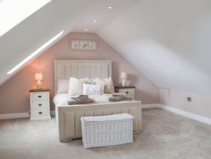 Une chambre avec un plafond en pente, un lit agrémenté de plusieurs oreillers, des tables de chevet avec des lampes, un panier en osier blanc et des œuvres d’art encadrées accrochées au mur.