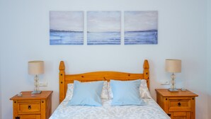 Ein Schlafzimmer mit einem hölzernen Kopfende, zwei Nachttischen jeweils mit einer Lampe, einem Bett mit gemusterten Betttextilien und drei an der Wand hängenden Bildern einer Küstenlandschaft.