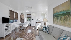 Una sala moderna con un sofá, una mesa de comedor y sillas. Hay una cocina con armarios y un refrigerador. Un cuadro adorna la pared.