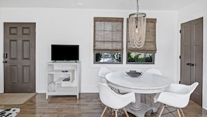 Un comedor con una mesa redonda, sillas blancas, un televisor sobre un estante blanco y una ventana con persianas de mimbre.
