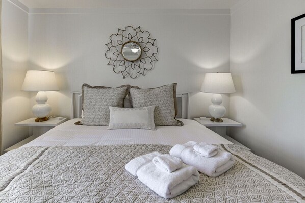 Una cama bien hecha con ropa de cama estampada y almohadas, dos lámparas de noche y un espejo decorativo en la pared.