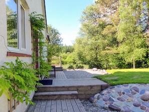 A wooden deck leading to a house with a stone wall and a garden with trees and a pond. - Bo Nan Taigh Cottage (Aberfoyle)