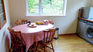 Ruokailutila, jossa on ruutukuviollinen pöytäliina, puiset tuolit, hedelmäkulho ja ikkuna, josta avautuu näkymä vehreään maisemaan.