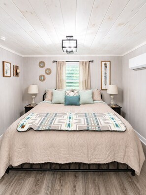 A bedroom with a bed, two bedside tables, a window with curtains, wall-mounted mirrors, and an air conditioning unit. - Escape to the Smokies! Tiny House Cabin-Special - 4 Night Min w/free cleaning! (Gatlinburg)