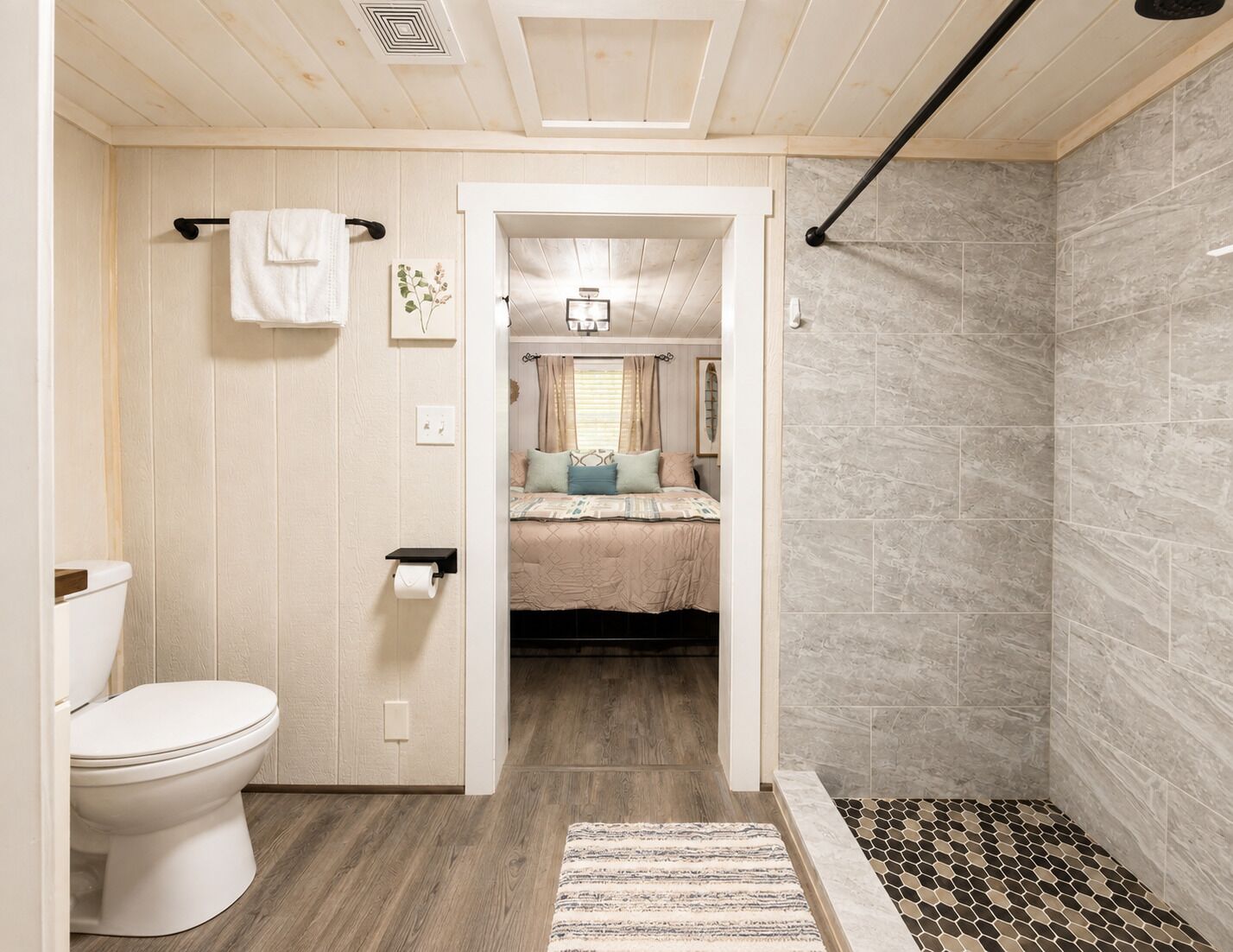 Un baño con inodoro, zona de ducha y una habitación visible con cama y ventana.